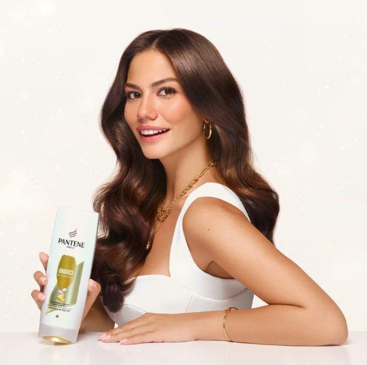 Frau mit glänzendem, braunem Haar hält Pantene Haarpflegeprodukt in der Hand. Perfekt gestyltes, gesund aussehendes Haar, betont durch goldene Accessoires und ein weißes Outfit. Haarpflege für Glanz und Stärke.