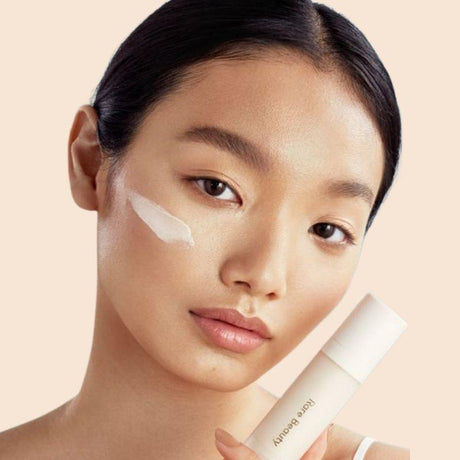 Nahaufnahme einer Frau mit makelloser Haut, die eine Foundation-Creme auf ihrer Wange trägt und ein Foundation-Produkt in der Hand hält.